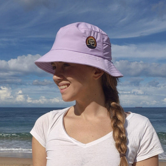 Woman wearing a lilac surf hat bucket style smiling with a beach and sand behind her braided hair