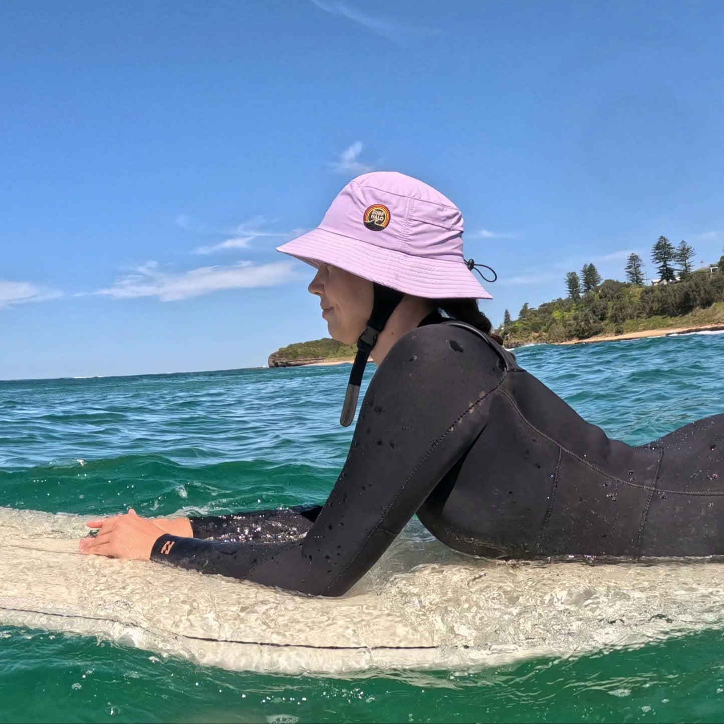 Person in a wetsuit and pink hat lying on a surfboard in the ocean with a clear blue sky.