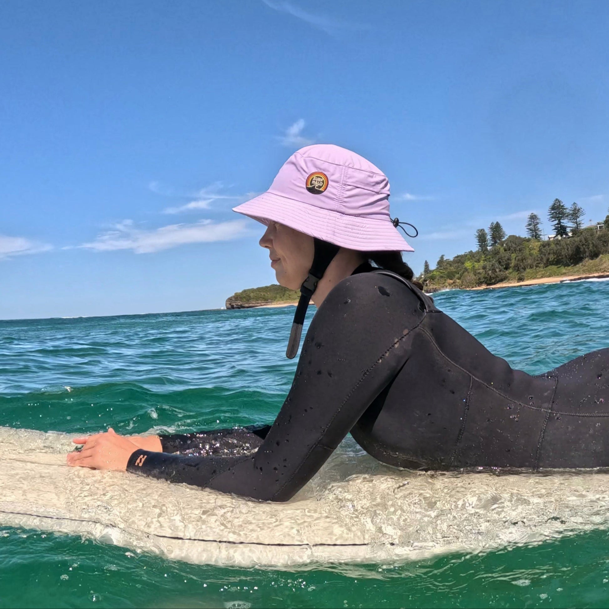 Person in a wetsuit and pink hat lying on a surfboard in the ocean with a clear blue sky.