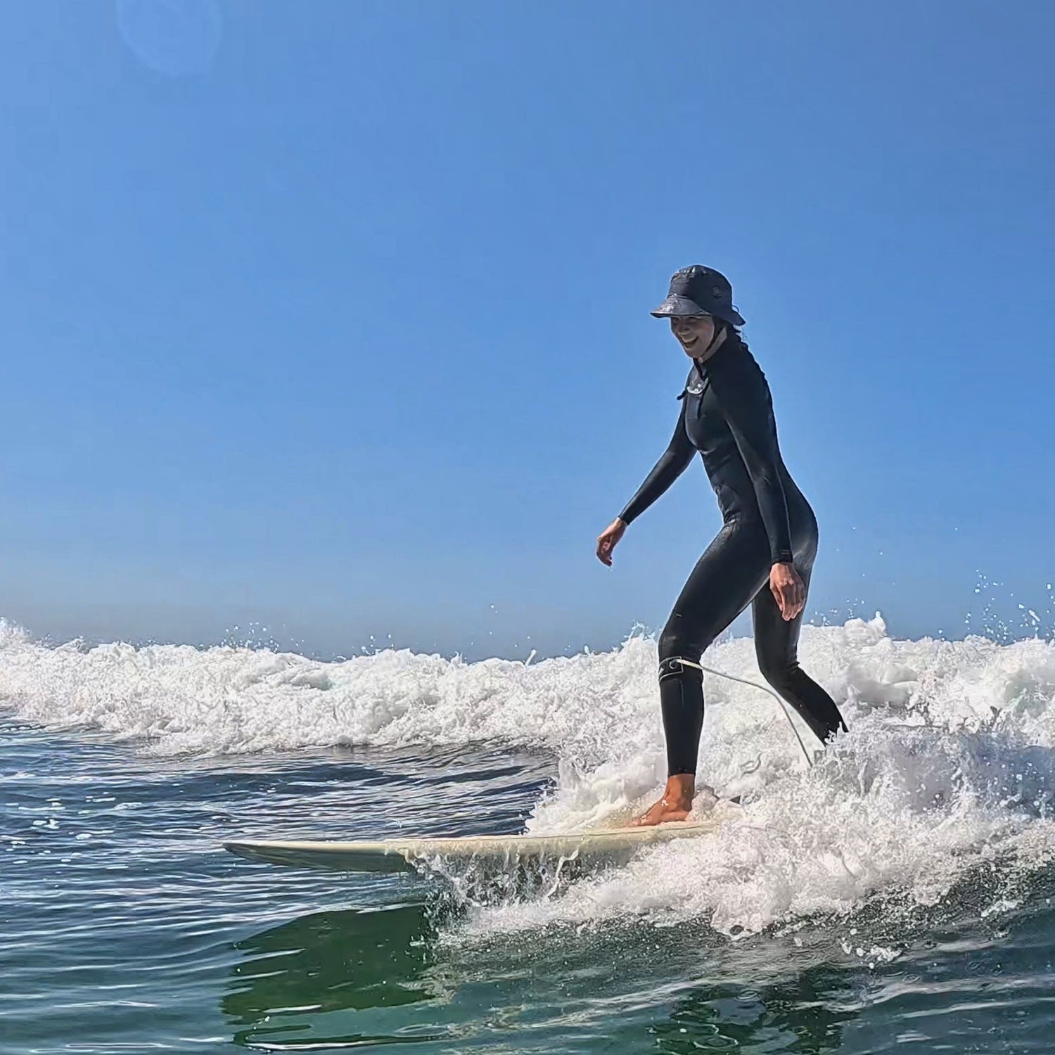 Surfer girl on a longboard on a small wave wearing a dark grey surf hat with a protective inner shell UPF50+ and a wetsuit