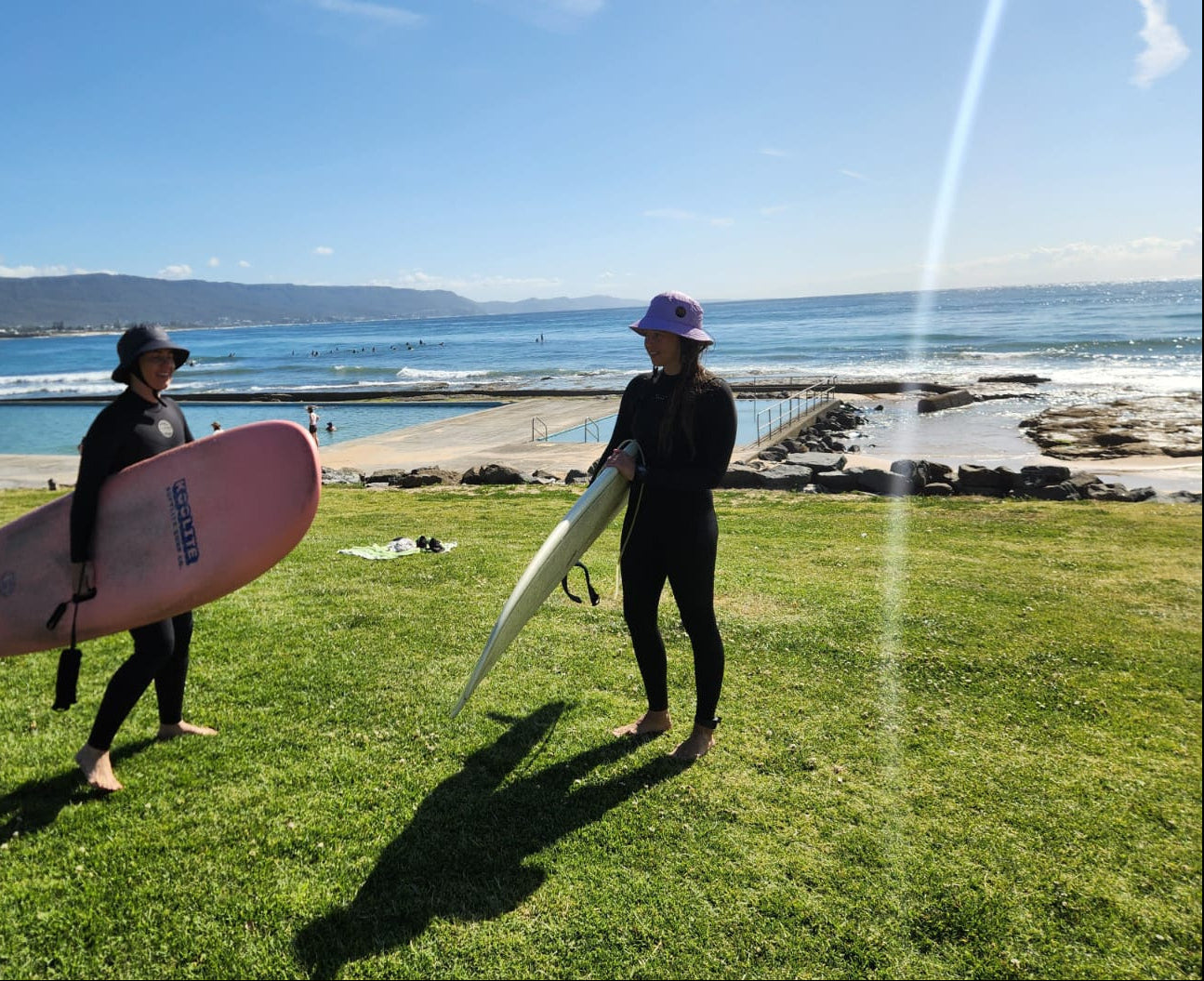 Two girsl with surfboards on a grassy area near a beach.