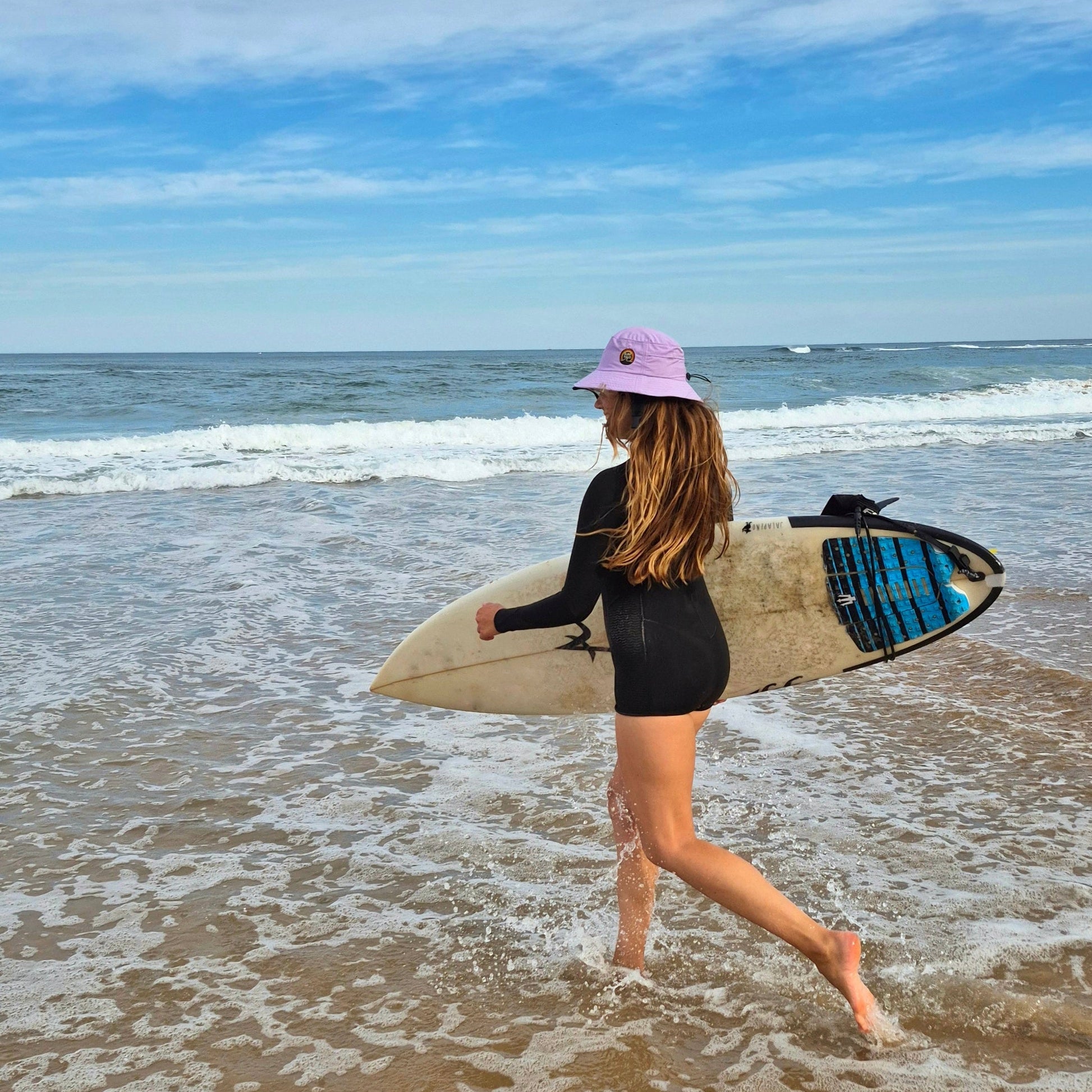 Woman walking on a beach with a surfboard, wearing a black swimsuit and pink hat.